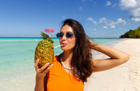 Satisfied lady in swimwear and sunglasses walking on sandy beach and enjoying cocktail drink from pineapple on tropical island - Powered by Adobe