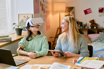 Obraz premium Mother sitting with daughter with prosthetic hand and wearing virtual reality headset in cozy bedroom illuminated by lamp, both engaged in learning and interaction