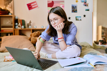 Caucasian teenager with prosthetic hand studying while using laptop in cozy bedroom environment,...