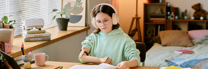 Child with prosthetic arm and headphones deeply engrossed in reading book while sitting at desk in well-decorated, cozy bedroom with plants and study materials around her