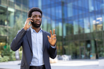 A handsome businessman gesturing while on his phone stands confidently in front of an office...