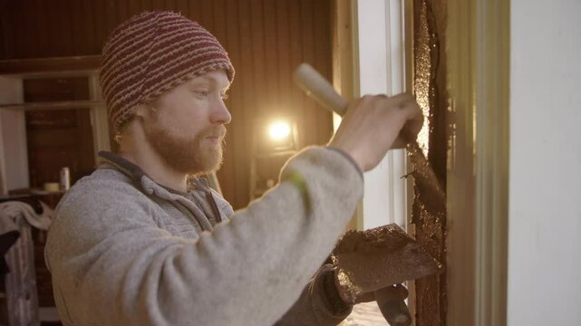 Nordic man backlit by floodlight applies clay plaster to house under renovation