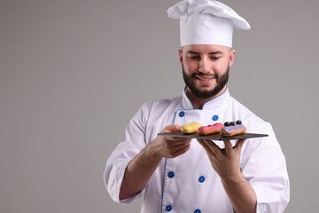 Happy confectioner in uniform holding delicious eclairs on light grey background