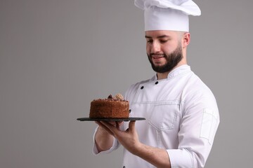 Happy confectioner in uniform holding delicious chocolate cake on light grey background