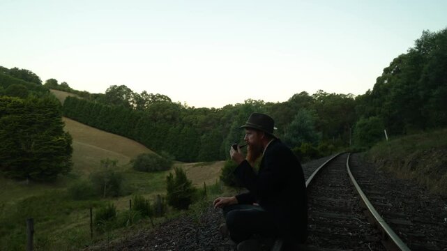 A traveling hobo smokes a tobacco pipe on a historic railway line while looking at rural farmland.