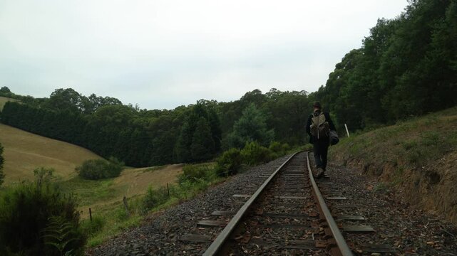 A traveling swagman walks a historic railway line along rural farmland in Australia.