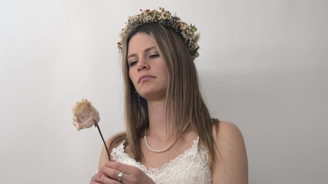 A bride, in a flowing white dress and floral crown, holds a single rose close to her. As she gazes down at the flower, her expression is tinged with sadness and disappointment.