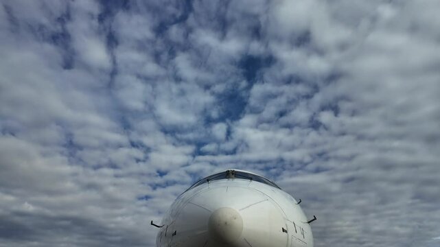 External frontal view of the nose of a white color fuselage jet airplane at the stand under a changing blue sky with some fluffy clouds. Low angle shot. Timelapse. 4K