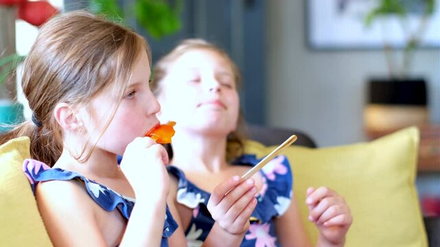 Caucasian fraternal twin girls enjoy orange ice pops, close-up side lighting