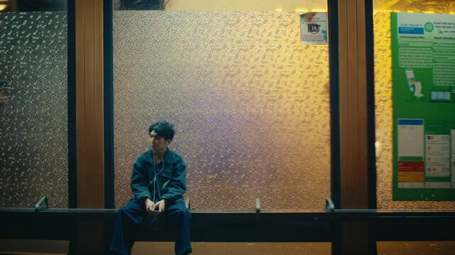 time lapse of lonely man sitting on bus stop station listening to music while people pedestrian walking in from of him , passing of time loneliness in the city , waiting concept