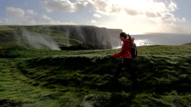 Asian woman hikes grassy Cliffs of Moher trail as Atlantic sea spray blows in wind