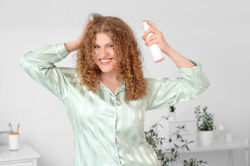 Naklejka premium Smiling young woman with curly hair applying hair spray for styling and care in pajamas, she is wearing pajamas, performing her morning beauty routine at home