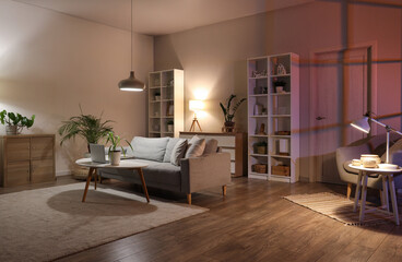 Sofa, shelving unit, houseplants and glowing lamps in interior of living room