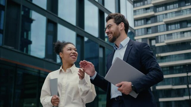 Two multiracial business people colleagues walking in city smiling friendly happy talking African American businesswoman woman female with Caucasian man businessman male work discuss holding folders