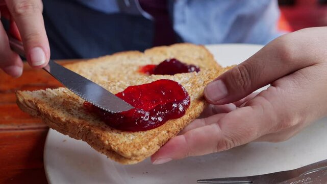 Closeup: European woman's hands spread blobs of red jam evenly across dry toast using stainless steel butter knife