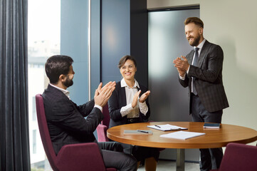 Team of happy business people sitting together around office table, making applauds and congratulating colleague with promotion, having corporate meeting and discussion. Business success