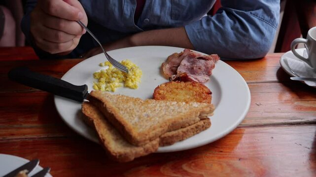 Eat scrambled eggs on breakfast plate with a fork, only hands visible. Toast, bacon and hashbrown too
