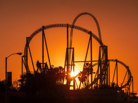 Knott's Berry Farm amusement theme park at sunset, sun peeking through roller coaster thrill rides silhouettes.
