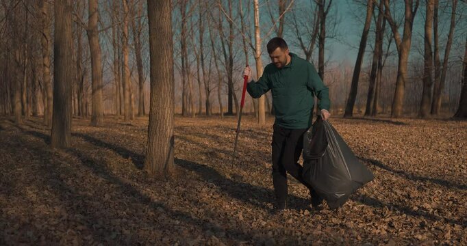 Man collecting plastic waste using a trash picker in a young forest during day time, blue sky, full body shot