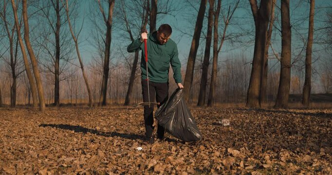 Man collecting plastic waste using a trash picker in a young forest during day time, blue sky, full body shot