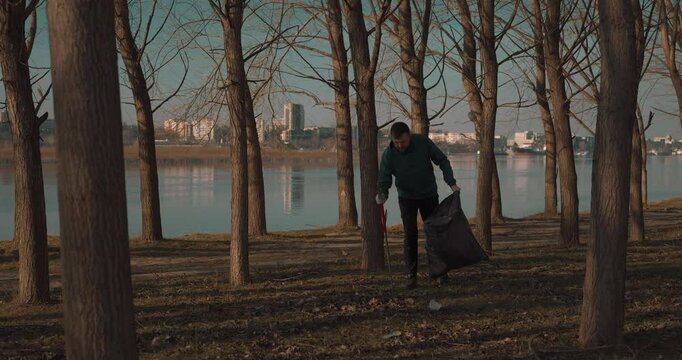 Man collecting plastic waste using a trash picker in a young forest near a river, city skyline in the background, blue sky