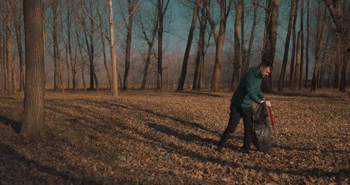 Man collecting plastic waste using a trash picker in a young forest during day time, blue sky, full body shot