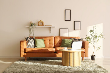 Interior of living room with houseplant, stylish leather sofa and laptop on table near white wall
