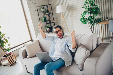 Young man celebrating success on laptop while sitting on couch in modern living room