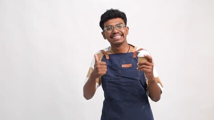 A cheerful young male barista wearing a navy blue apron and striped casual t-shirt is smiling while holding a takeaway coffee cup in one hand and pointing to aside