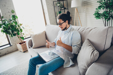 Young man giving thumbs up while working on a laptop on a cozy sofa in a bright living room.