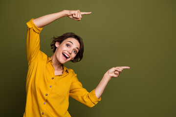 Cheerful young woman in a yellow shirt pointing joyfully with both hands against a solid green...