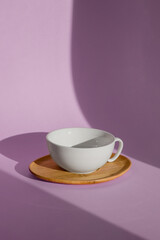 Fototapeta premium A white cup on a wooden saucer is depicted on a purple background. The concept of tableware for tea and coffee, cutlery.