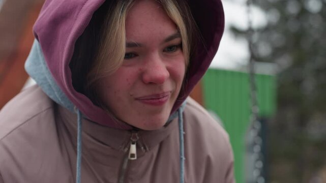 Close-up of young lady bent forward in winter outfit, feeling discomfort, hooded jacket covers head, face slightly strained, with blurred background featuring green structures and outdoor elements