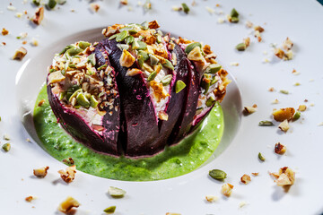 Beetroot with cottage cheese cream, nuts and pumpkin seeds on green spinach sauce is served on a white plate. Restaurant serving of a healthy dish. Close-up.