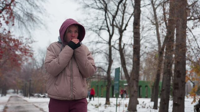 Calm woman in winter outfit strolling alone through a snow-covered park, bringing hands close to mouth for warmth, bare trees, scattered snow, and blurred figures in the background