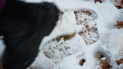Close-up of black boot jumping on icy ground, leaving distinct shoe imprint in fresh snow, scattered dry leaves add texture to the frozen landscape
