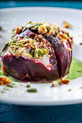 Beetroot with cottage cheese cream, nuts and pumpkin seeds on green spinach sauce is served on a white plate. Restaurant serving of a healthy dish. Close-up.
