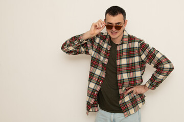 A man confidently poses while wearing a plaid shirt and sunglasses. He stands with one hand on his...