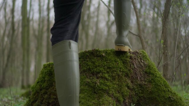 A person in rubber boots is standing on a mossy tree stump while surrounded by lush greenery in a forest