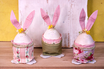 three handmade Easter bunnies made of eggs with pink ears stand against a yellow wall