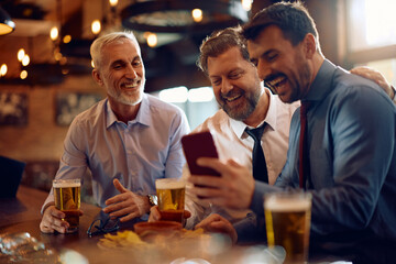 Cheerful businessmen using mobile phone while gathering for beer in bar.