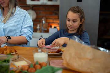 Young girl carefully placing ham on bread, making a sandwich during breakfast preparation in the kitchen, while her mother prepares other food items on the table