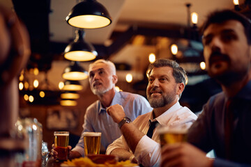Happy business colleagues watching sports game on TV while drinking beer in bar.