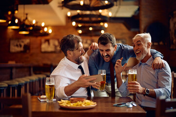Group of happy businessmen cheering while watching soccer match on mobile phone in pub.