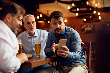 Group of businessmen watching sports match on smart phone in bar.