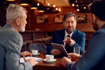 Mature entrepreneur communicating with coworkers while using touchpad on business meeting in cafe.