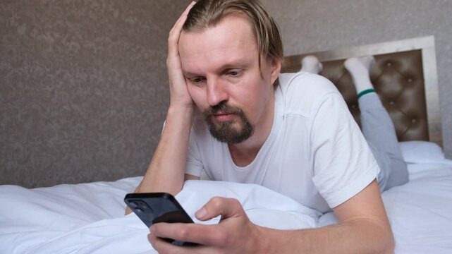 Young handsome attractive man with beard lying on bed with stomach down and using mobile phone to browse marketplace and talking to himself. Leisure of a slacker