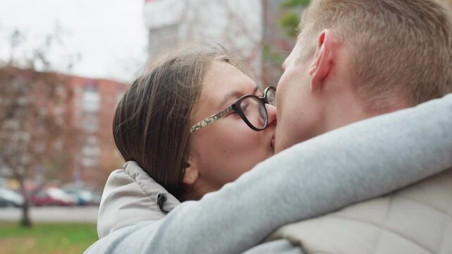 Close up of husband and wife kissing affectionately outdoors while holding each other warmly, sharing romantic moment in casual matching jackets with trees and blurred city background