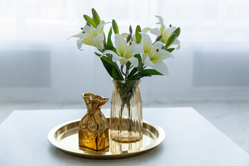Bouquet of white artificial lilies in a golden vase on a table on a light background.