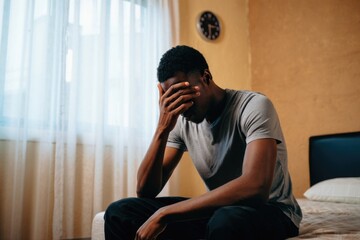 Fototapeta premium Stressed young african male sitting on bed in bedroom interior